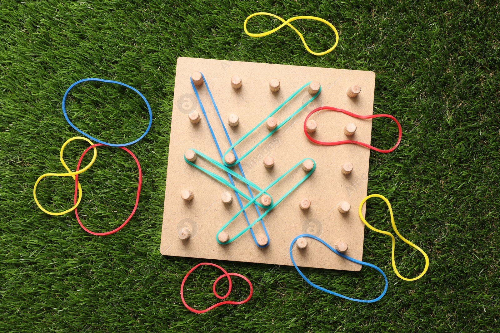 Photo of Wooden geoboard with rubber bands on artificial grass, flat lay. Educational toy for motor skills development