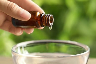 Photo of Woman pouring essential oil from glass bottle into bowl against blurred green background, closeup