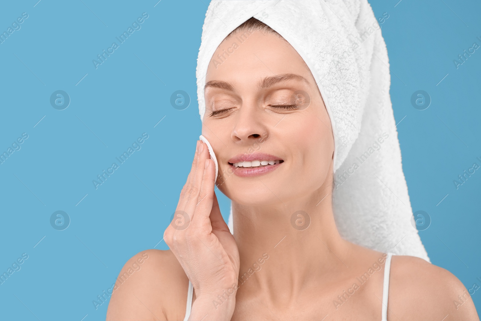 Photo of Beautiful woman in terry towel removing makeup with cotton pad on light blue background, space for text