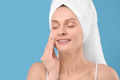 Beautiful woman in terry towel removing makeup with cotton pad on light blue background, space for text