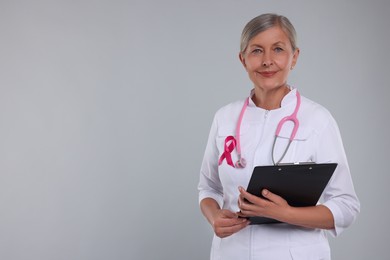 Photo of Doctor with pink ribbon, clipboard and stethoscope on light grey background, space for text. Breast cancer awareness