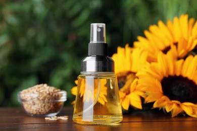 Photo of Spray bottle with cooking oil near sunflower seeds and flowers on wooden table against blurred green background