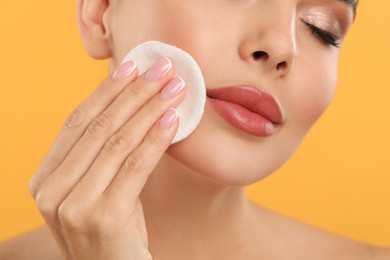Beautiful woman removing makeup with cotton pad on orange background, closeup