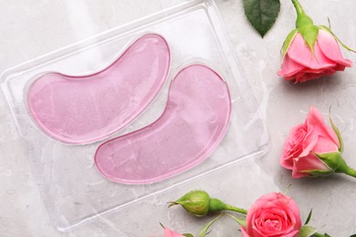 Package of under eye patches and rose flowers on light grey table, flat lay. Cosmetic product