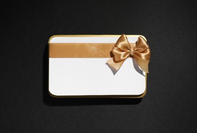 Photo of Blank gift card with golden bow on black background, top view