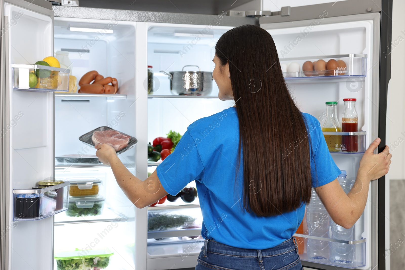 Photo of Young woman taking pack of meat out of refrigerator indoors, back view