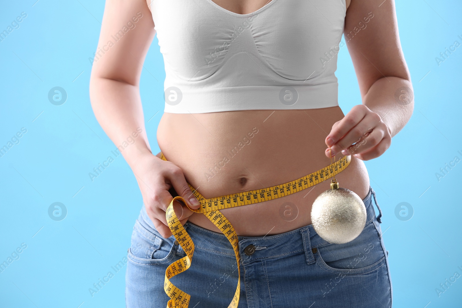 Photo of Woman with Christmas ball and measuring tape on light blue background, closeup. Overweight problem after New Year party
