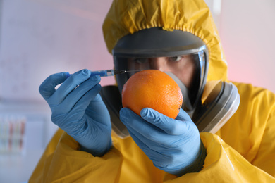 Scientist in chemical protective suit injecting orange at laboratory, focus on fruit