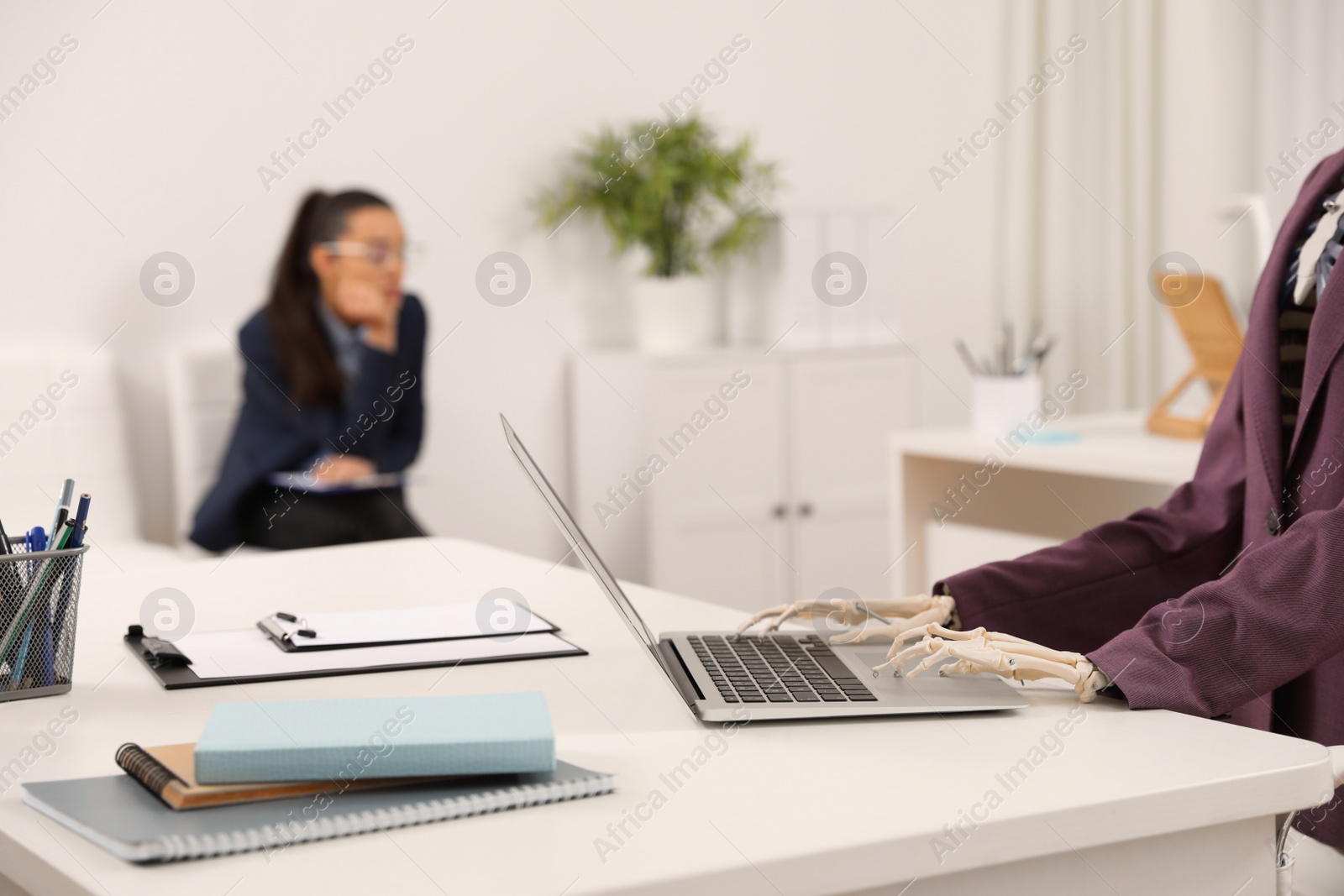 Photo of Human skeleton in suit using laptop at table and woman waiting on chair in office, closeup