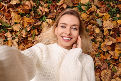 Smiling woman lying among autumn leaves and taking selfie outdoors, top view