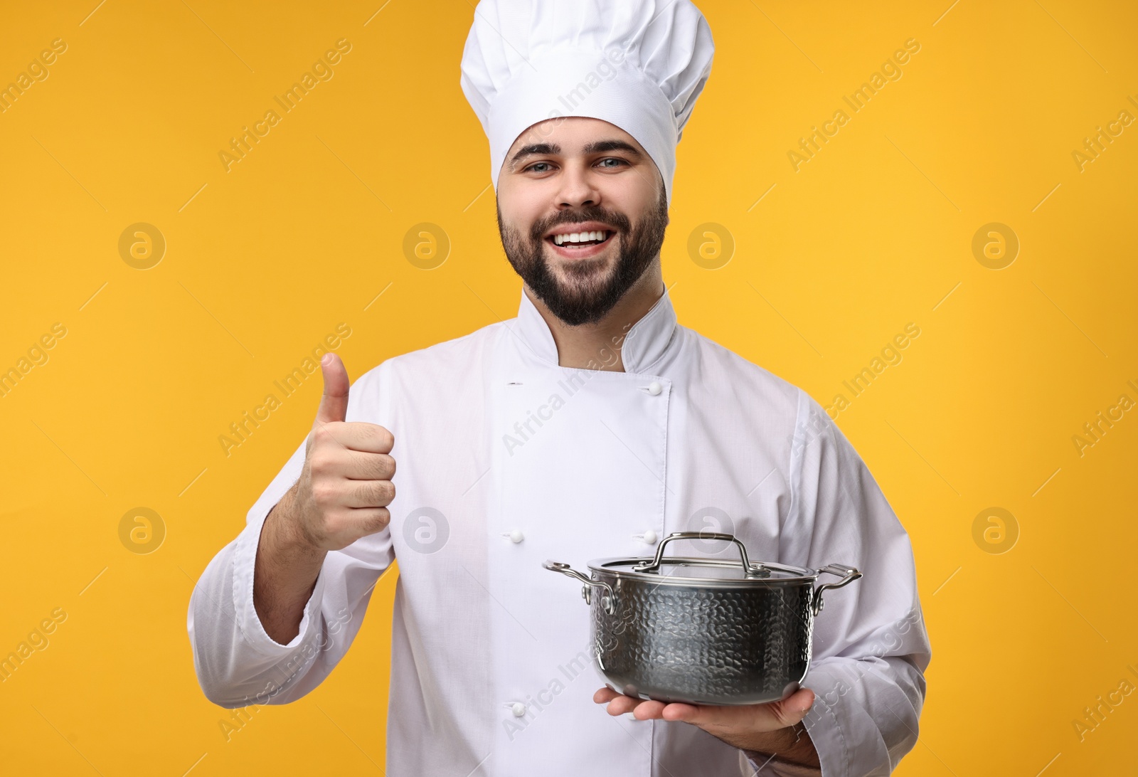 Photo of Happy young chef in uniform holding cooking pot and showing thumb up on orange background