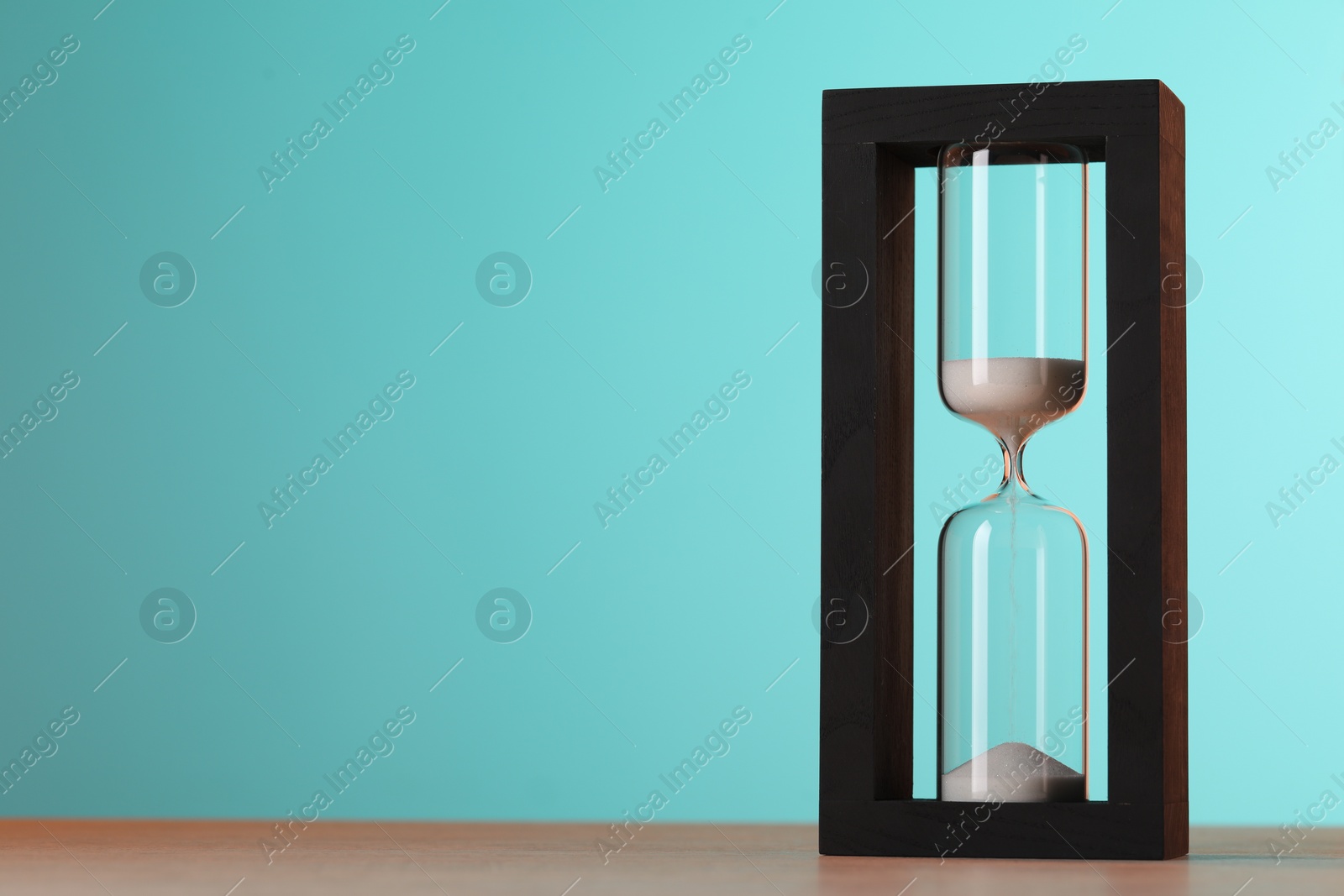 Photo of Hourglass with flowing sand on table against light blue background, space for text