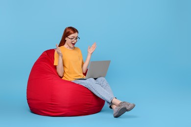 Surprised young woman with laptop sitting on beanbag chair against light blue background, space for text