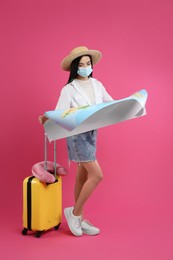 Female tourist in medical mask with map and suitcase on pink background. Travelling during coronavirus pandemic
