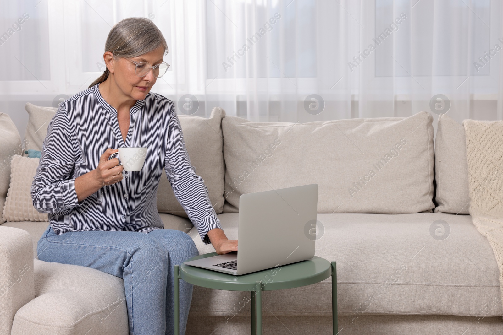 Photo of Beautiful senior woman with cup of drink using laptop at home