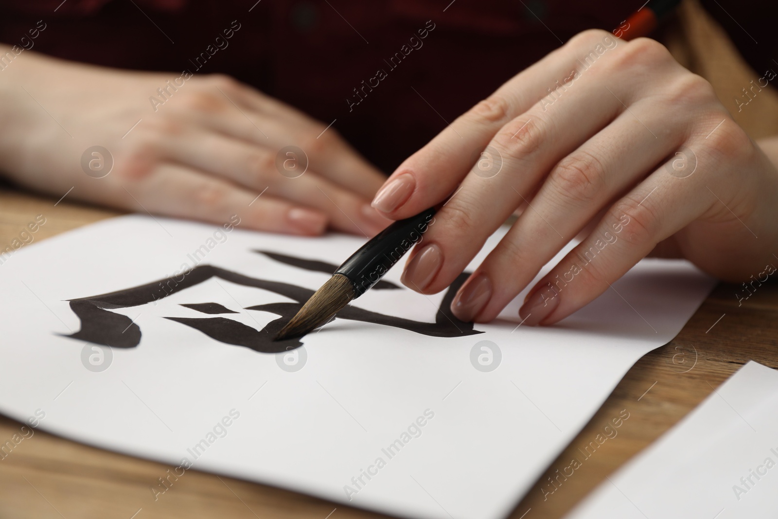 Photo of Calligraphy. Woman with brush writing hieroglyphs on paper at wooden table, closeup