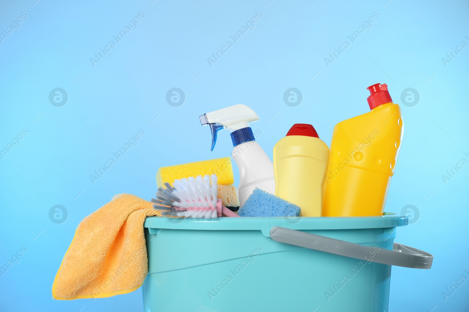 Photo of Bucket with different cleaning products and supplies on light blue background, closeup