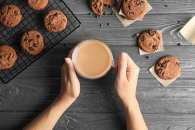 Photo of Woman holding cup of coffee near tasty chocolate chip cookies on wooden background, top view
