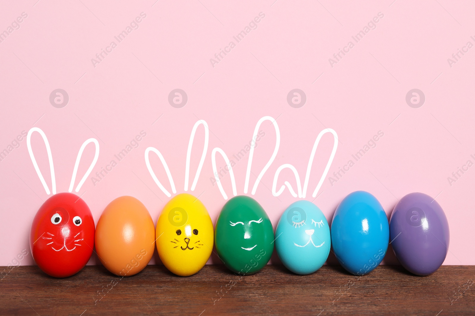 Image of Several eggs with drawn faces and ears as Easter bunnies among others on wooden table against pink background