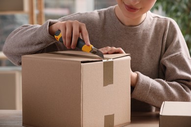 Young woman using utility knife to open parcel at wooden table indoors, closeup