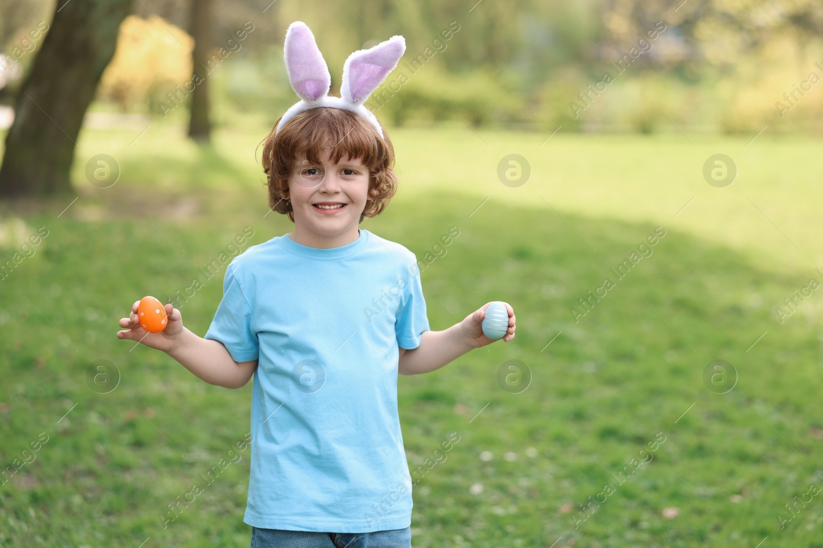 Photo of Easter celebration. Cute little boy in bunny ears holding painted eggs outdoors, space for text