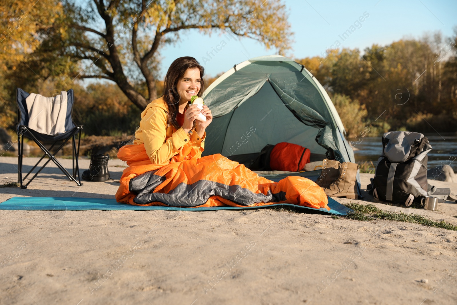 Photo of Young woman eating sandwich in sleeping bag near camping tent outdoors