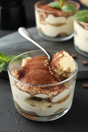 Photo of Delicious tiramisu in glasses and spoon on black table, closeup