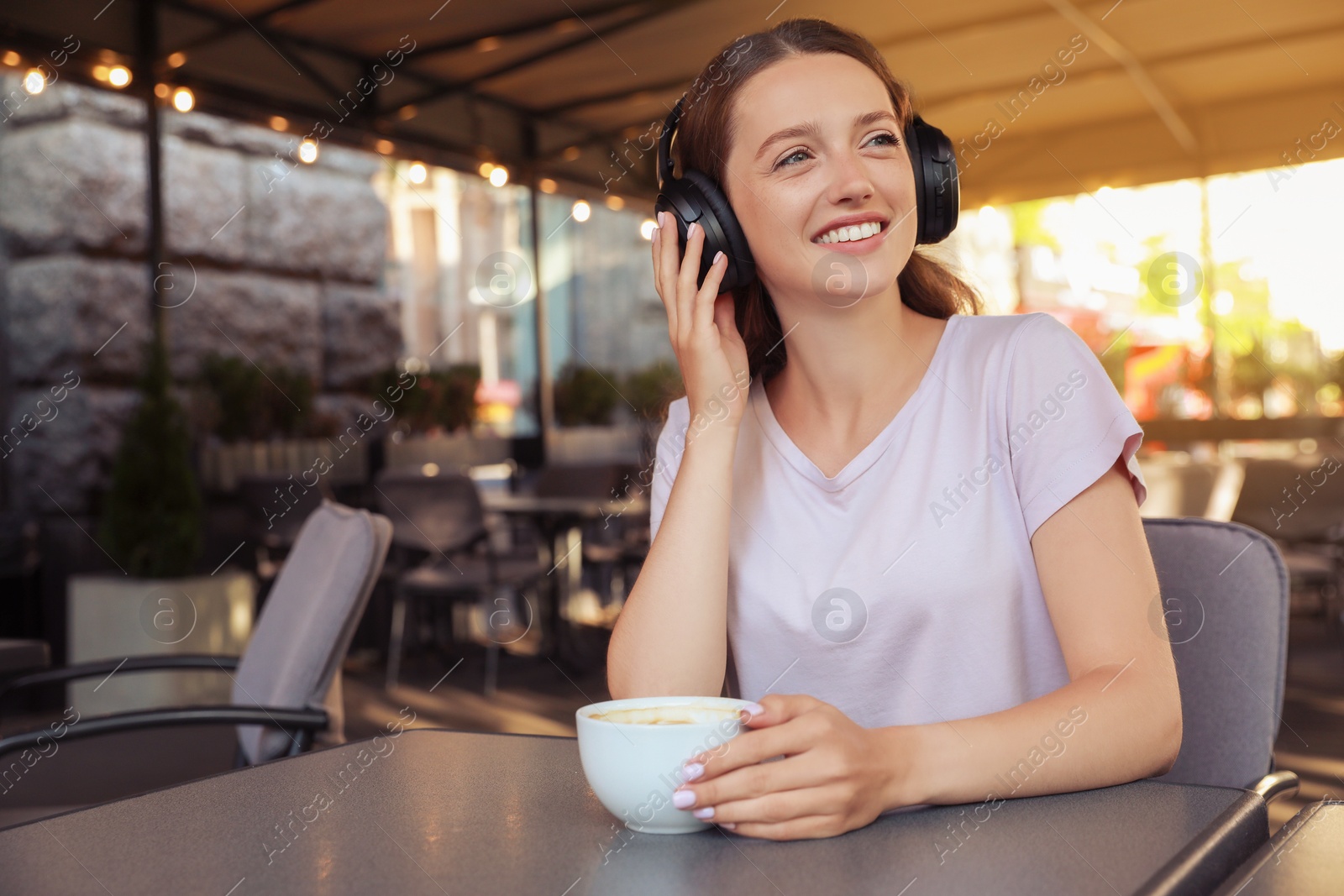 Photo of Smiling woman in headphones with cup of coffee in outdoor cafe. Space for text