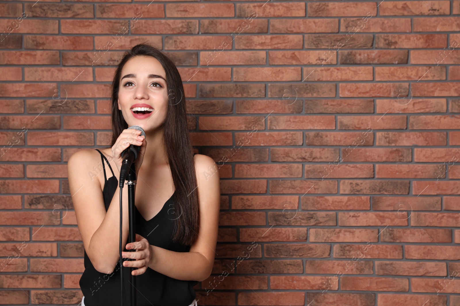 Photo of Young stylish woman singing in microphone near brick wall. Space for text