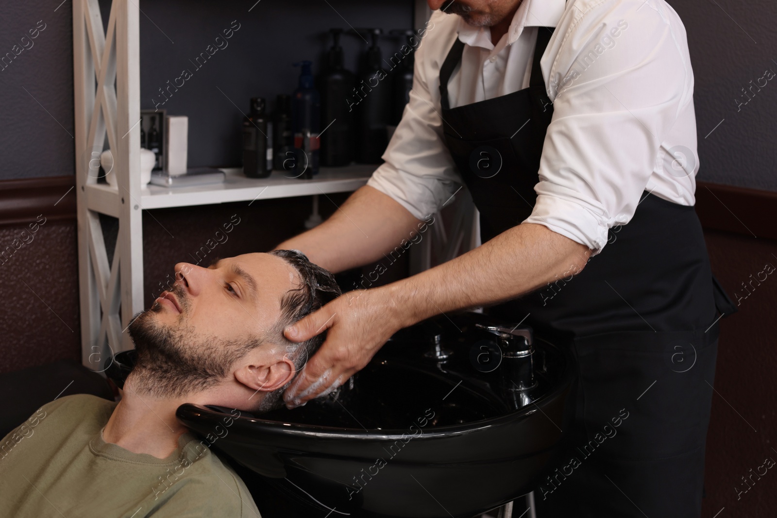 Photo of Professional hairdresser washing man's hair at sink in barbershop