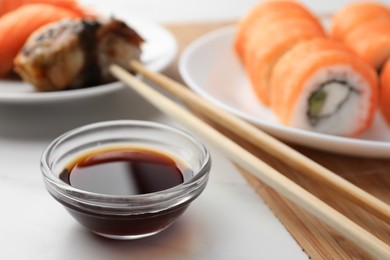 Bowl of tasty soy sauce, chopsticks and different types of sushi on white table, closeup