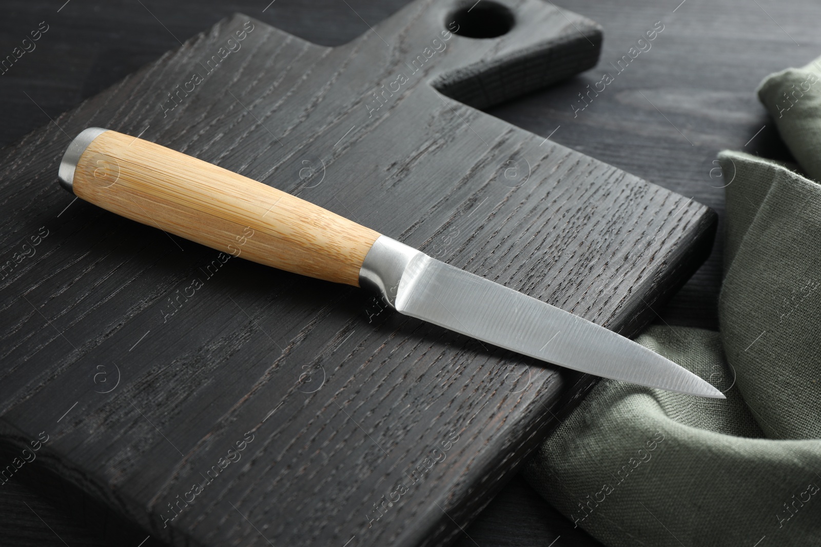 Photo of One sharp knife, cloth and board on black wooden table, closeup