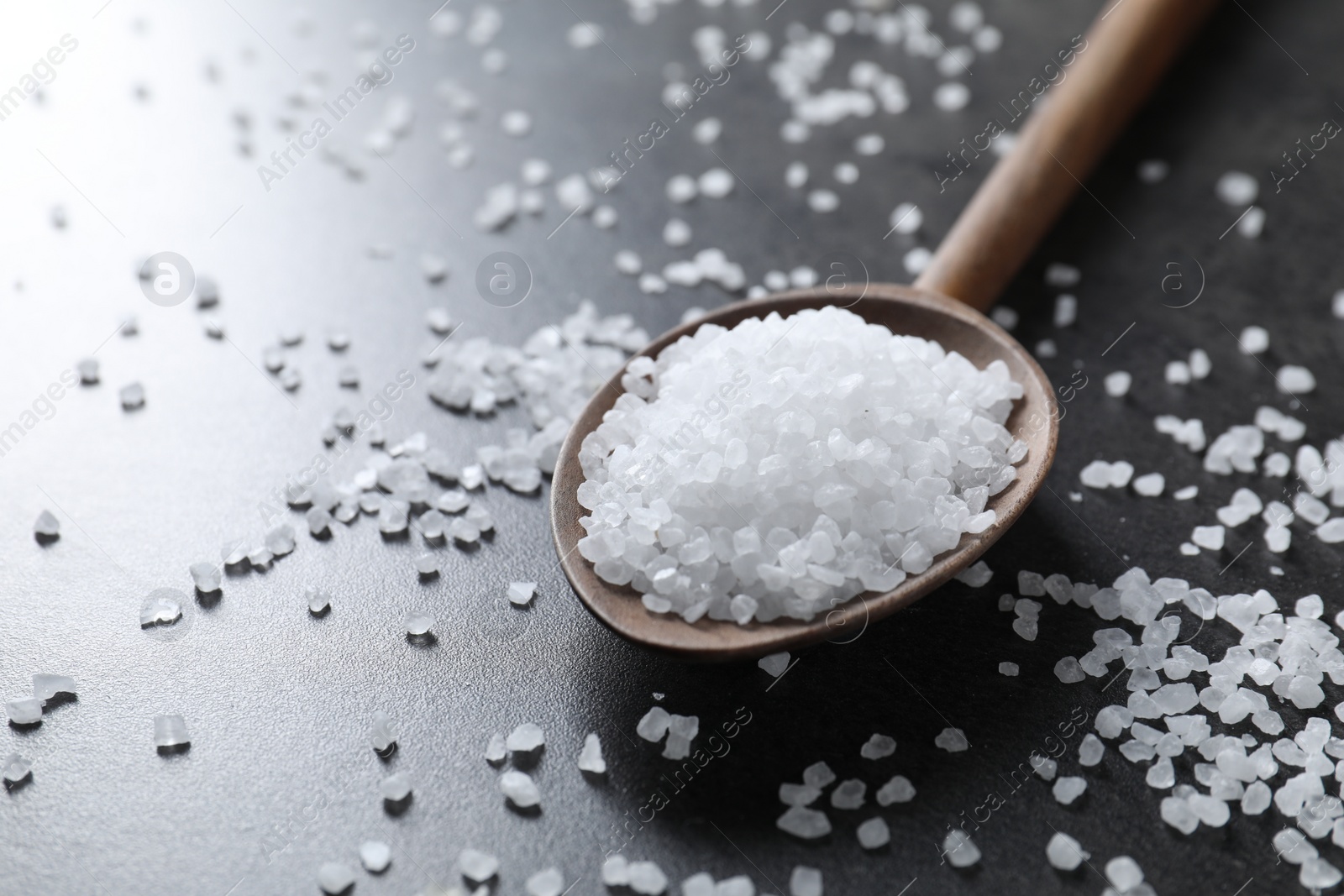 Photo of Natural salt and wooden spoon on black table, closeup. Space for text