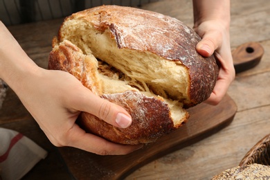 Woman breaking freshly baked bread at wooden table, closeup