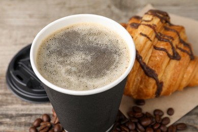 Photo of Coffee to go. Paper cup with tasty drink, beans and croissant on wooden table, closeup