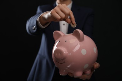 Woman putting coin into piggy bank on black background, closeup