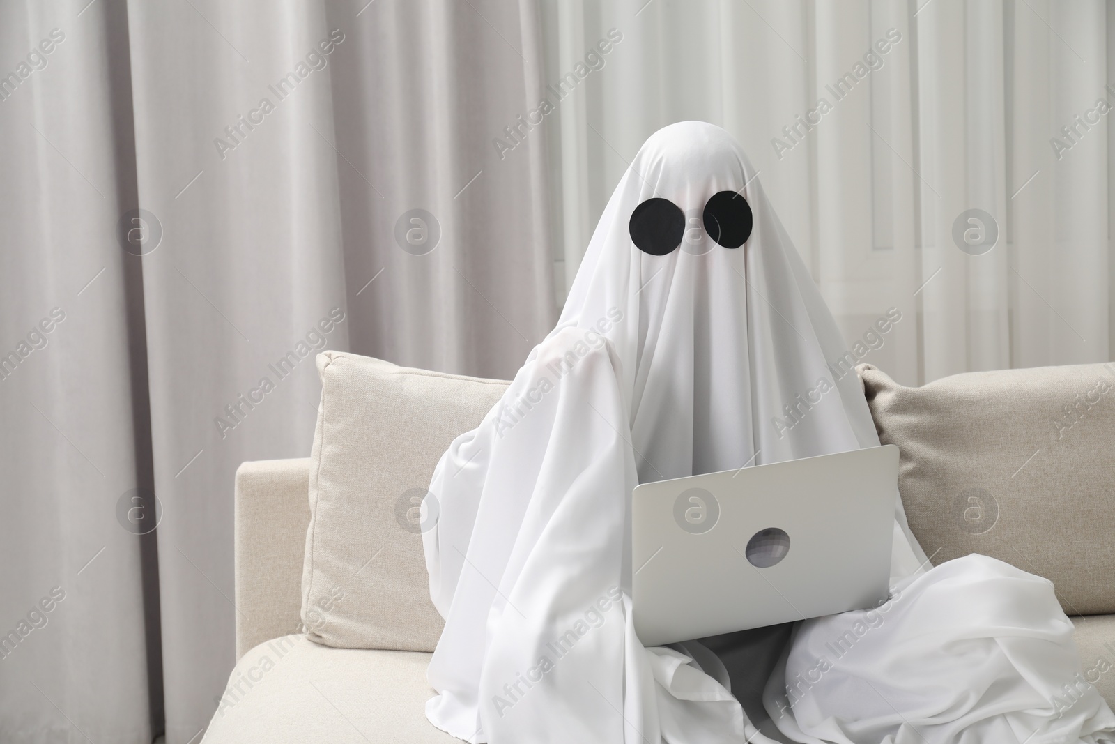 Photo of Creepy ghost. Person covered with white sheet using laptop on sofa at home