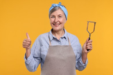 Photo of Happy housewife with potato masher showing thumbs up on orange background
