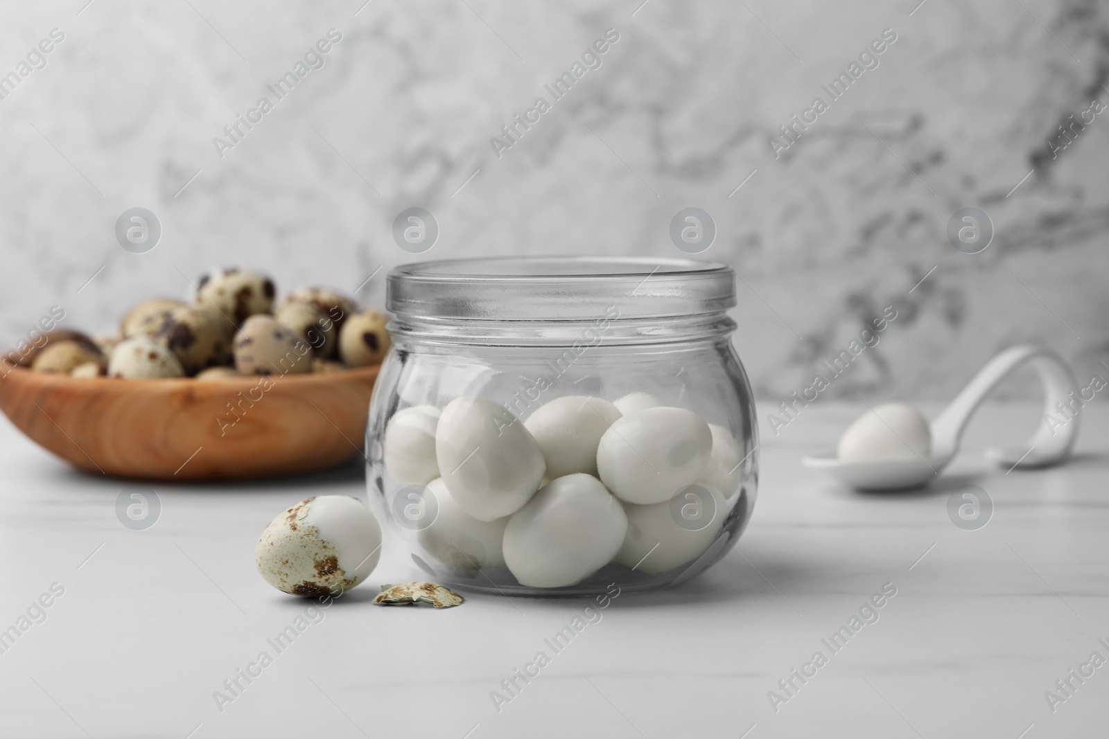 Photo of Glass jar with peeled boiled quail eggs and another one partly in shell on white table