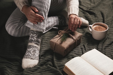 Photo of Woman in knitted socks sitting on warm plaid and unpacking gift box, closeup