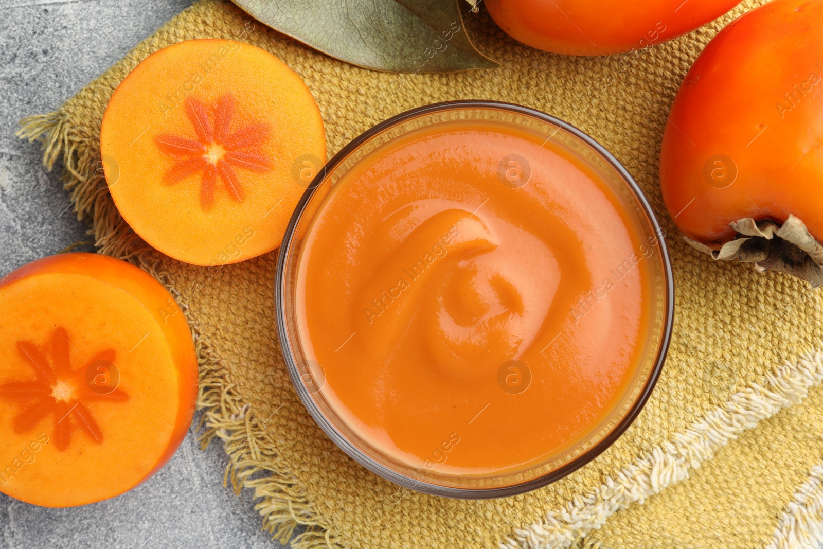 Photo of Delicious persimmon jam in glass bowl and fresh fruits on gray textured table, flat lay