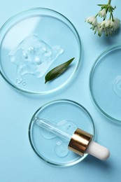 Photo of Petri dishes with samples of cosmetic oil, pipette and flowers on light blue background, flat lay