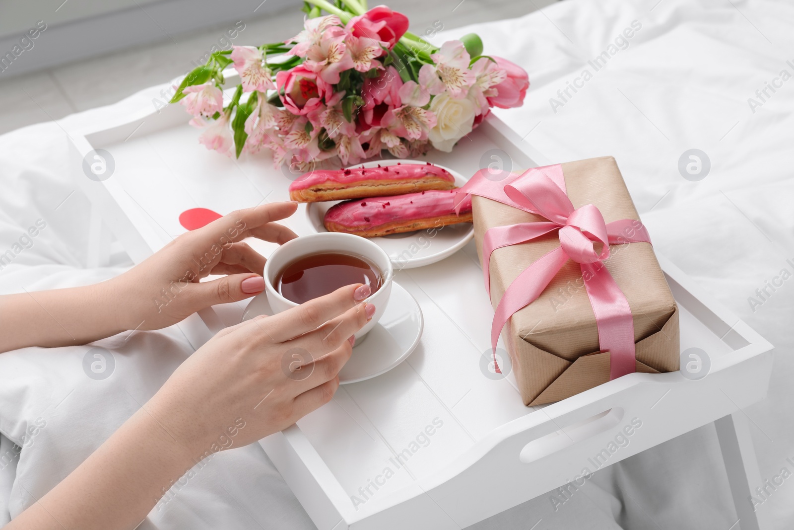 Photo of Tasty breakfast served in bed. Woman with tea, eclairs, gift box and flowers at home, closeup