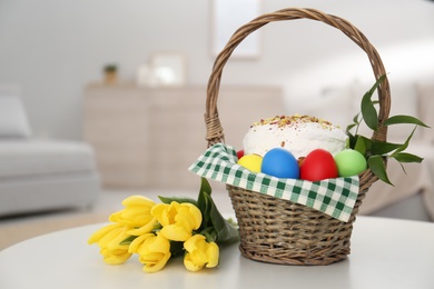 Basket with delicious Easter cake, dyed eggs and flowers on white table indoors. Space for text