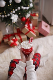Photo of Woman with cupdelicious hot drink near Christmas tree at home, closeup