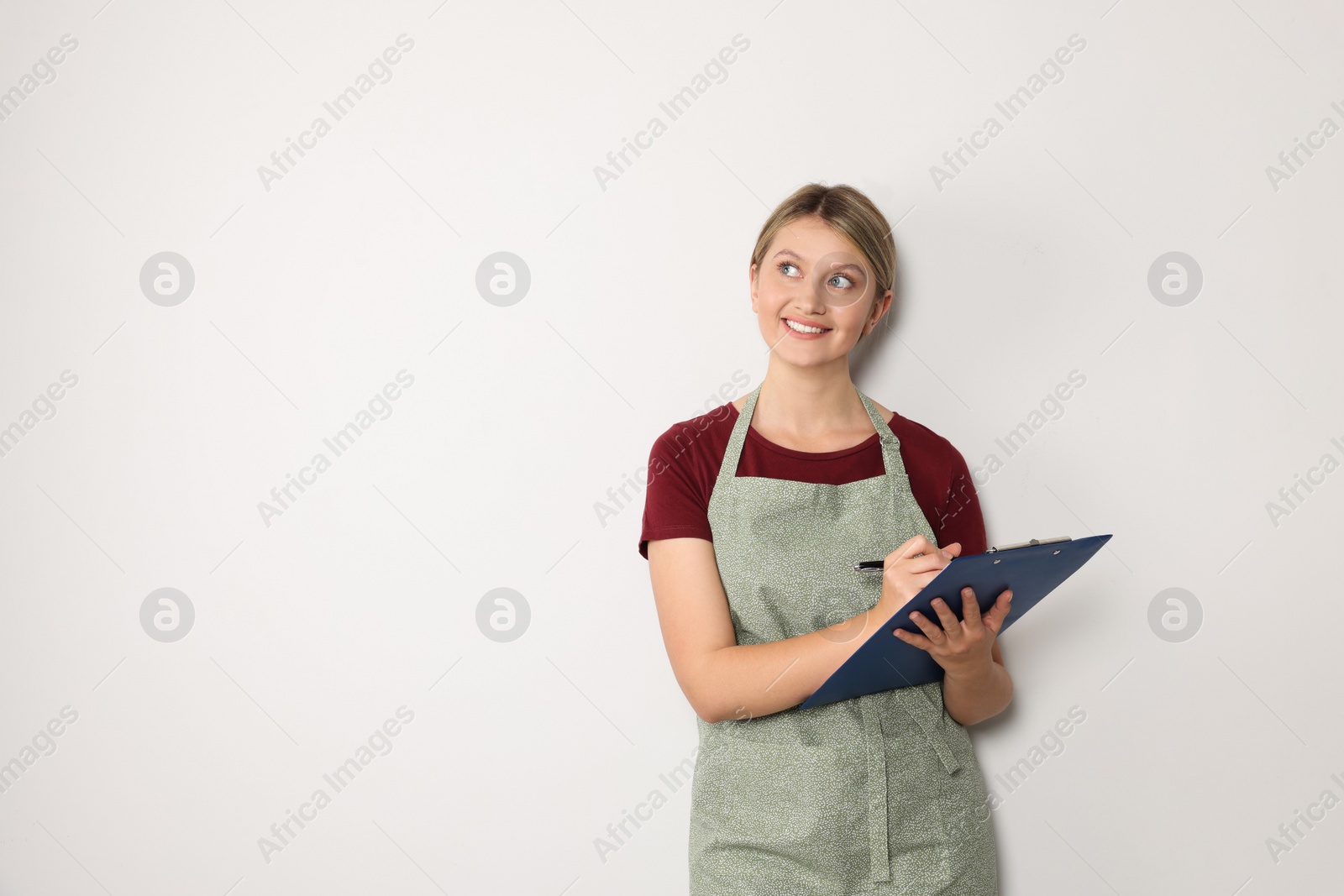 Photo of Beautiful young woman in clean apron with clipboard on light grey background. Space for text