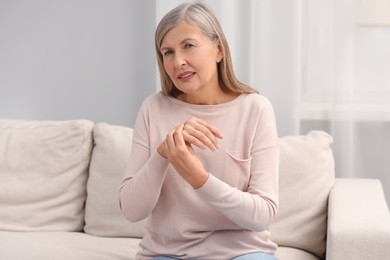 Photo of Mature woman suffering from pain in hand on sofa at home. Rheumatism symptom