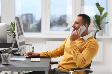 Young man talking on smartphone while working with computer at table in office