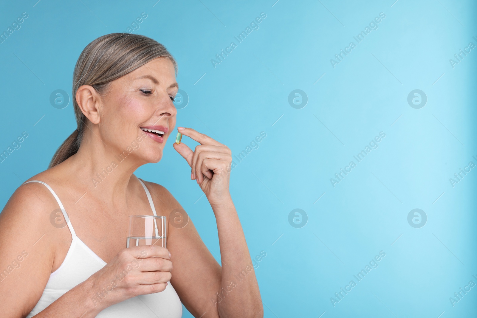 Photo of Beautiful woman with glass of water taking vitamin capsule on light blue background, space for text