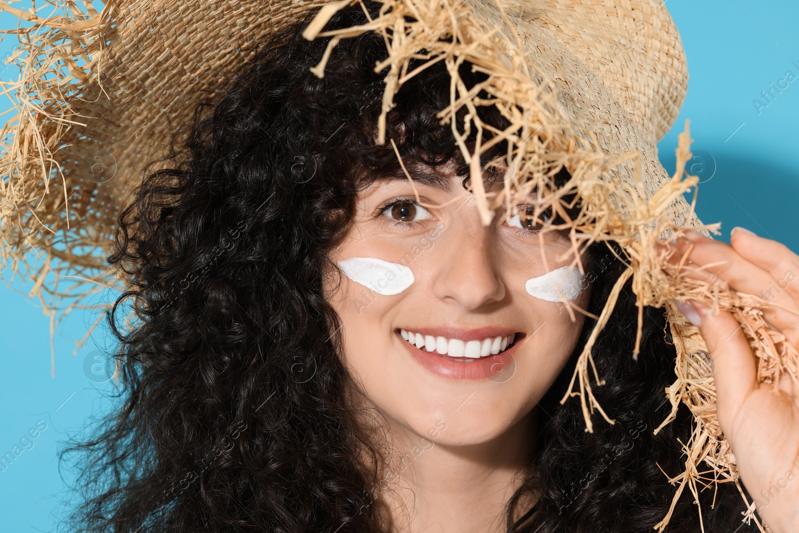 Photo of Beautiful young woman in straw hat with sun protection cream on her face against light blue background